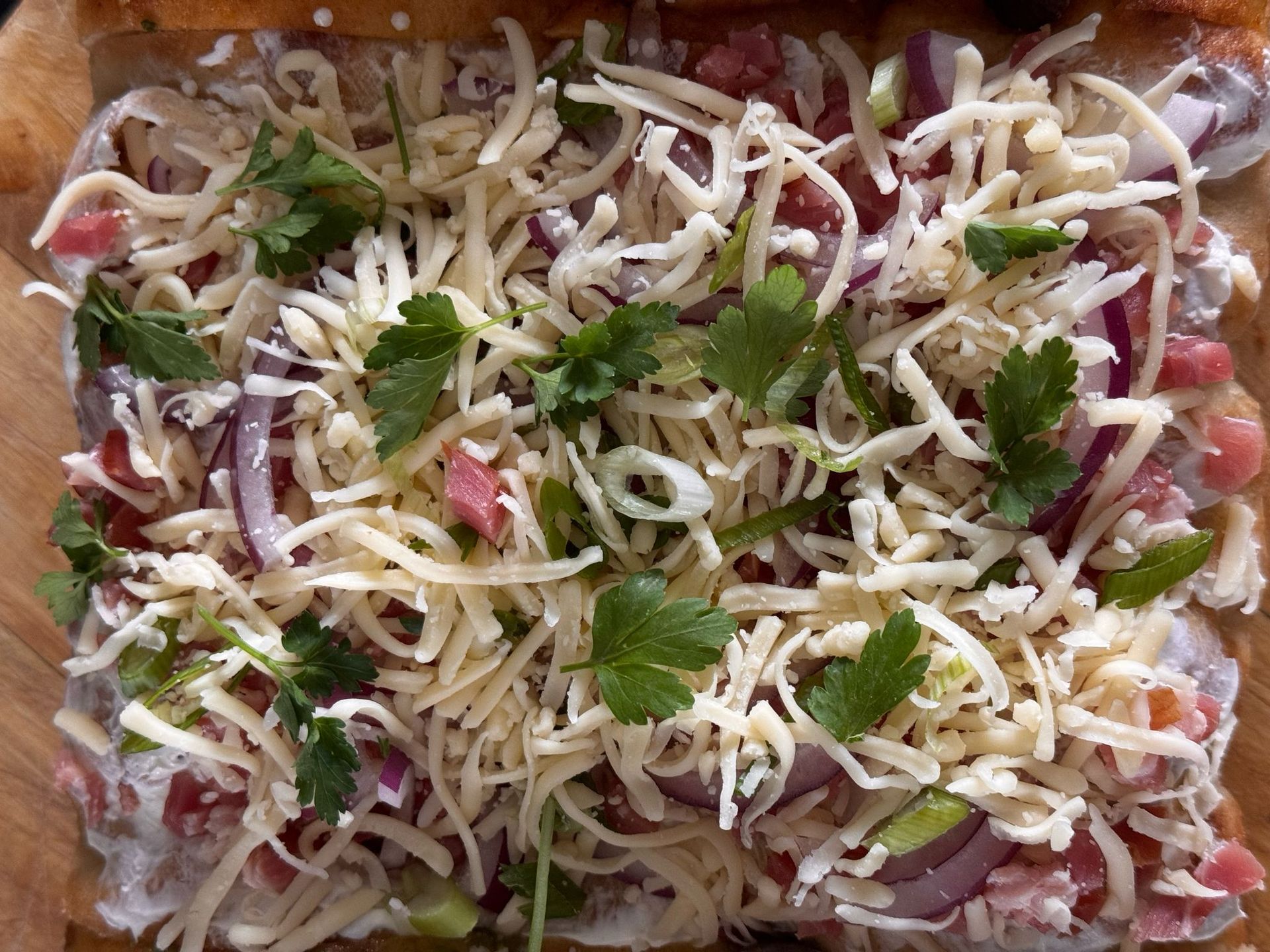 Ongebakken platbrood belegd met geraspte kaas, rode ui, blokjes ham of spek en verse koriander, geserveerd op een houten plank.