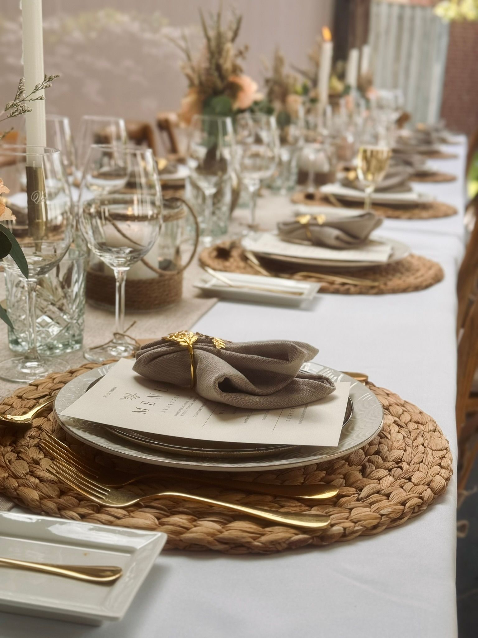Een lange eettafel gedekt met geweven onderborden, witte borden, gouden bestek, servetten met ringen en kaarsen voor een feestelijke gelegenheid.