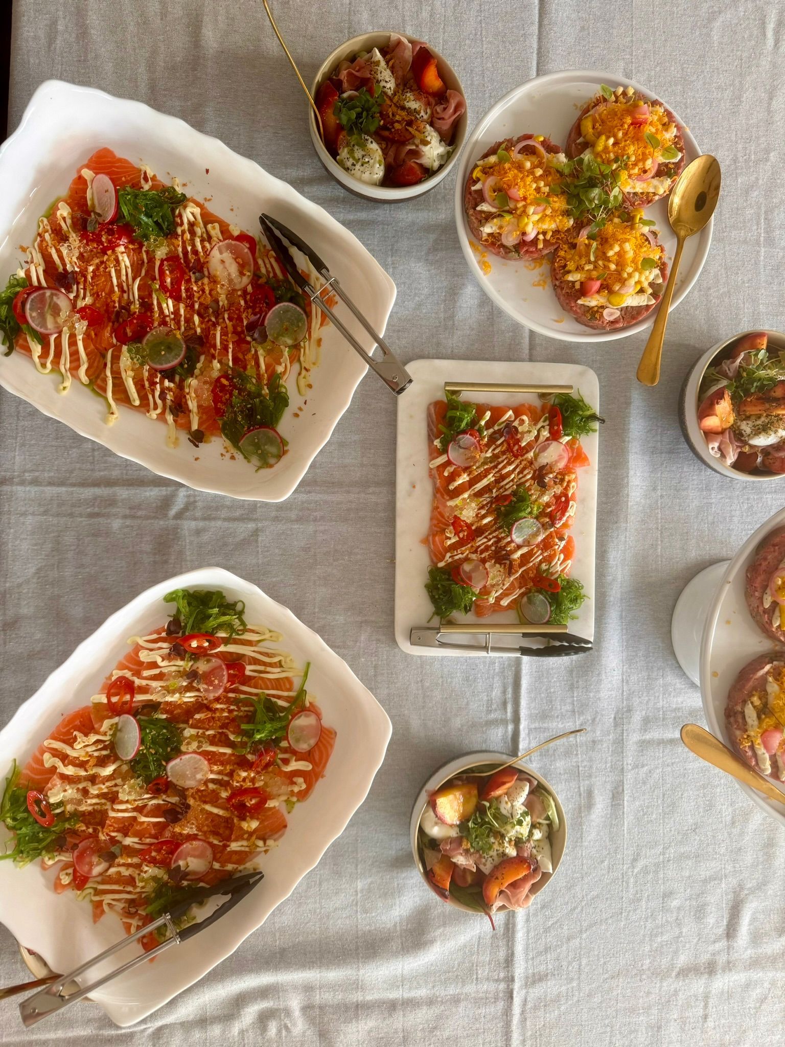 Bovenaanzicht van een tafel gedekt met diverse schalen zalmcarpaccio, kleine schaaltjes met bijgerechten en geroosterd brood.