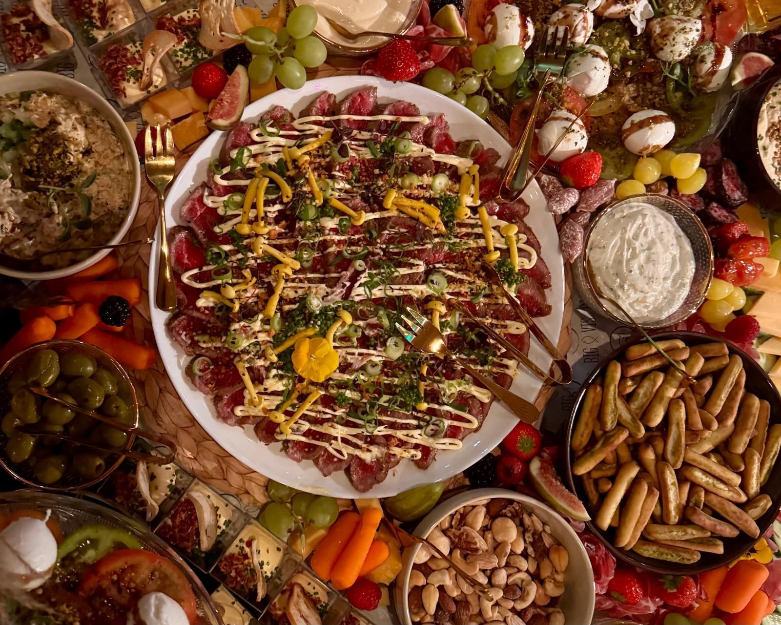 Een grote, ronde schaal met carpaccio van rundvlees, overgoten met sauzen, omringd door diverse hapjes op een borrelplank.