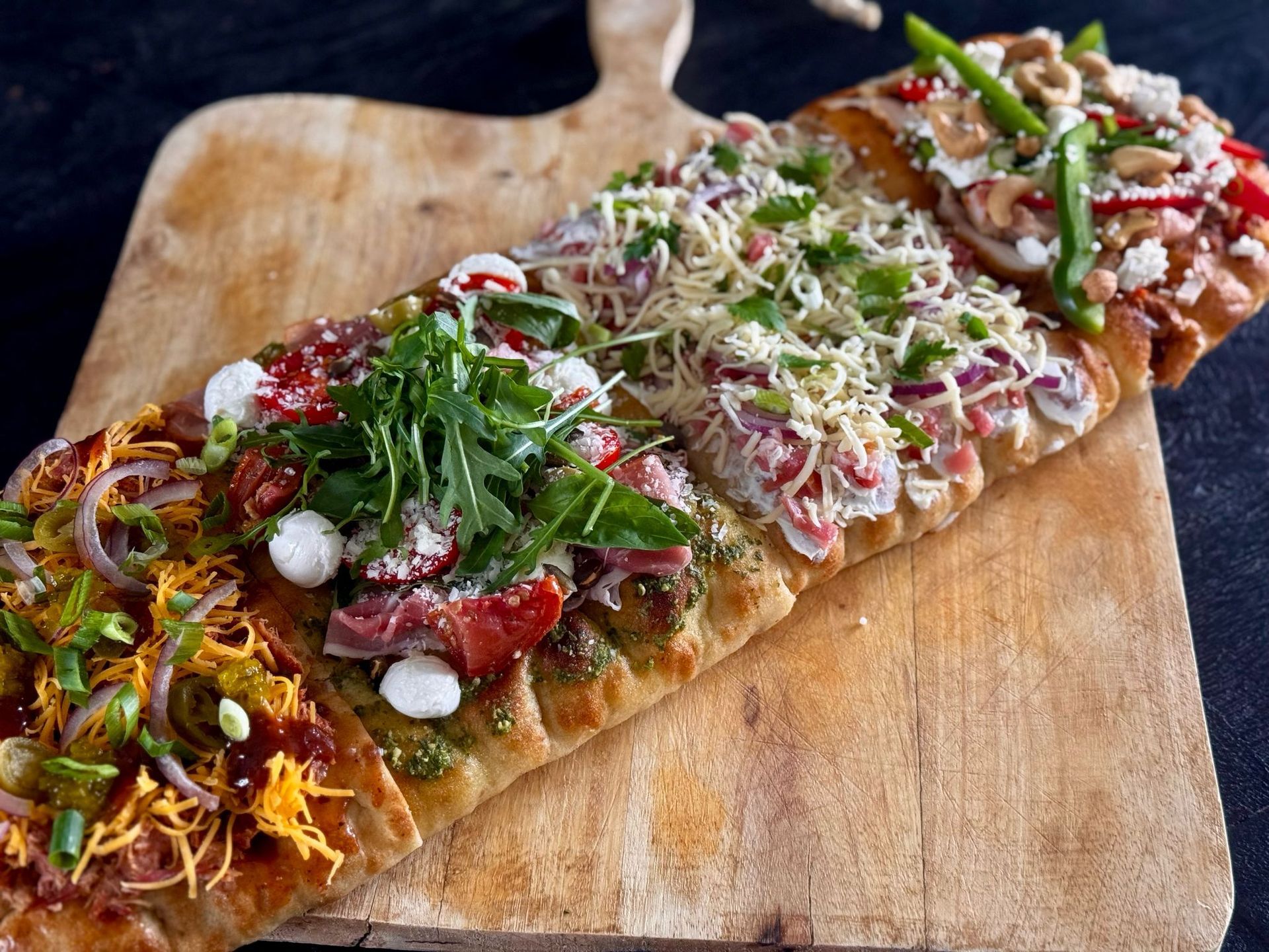 Een lange, rechthoekige platbroodpizza, in vier verschillende vakken met diverse toppings gesneden en geserveerd op een houten plank.