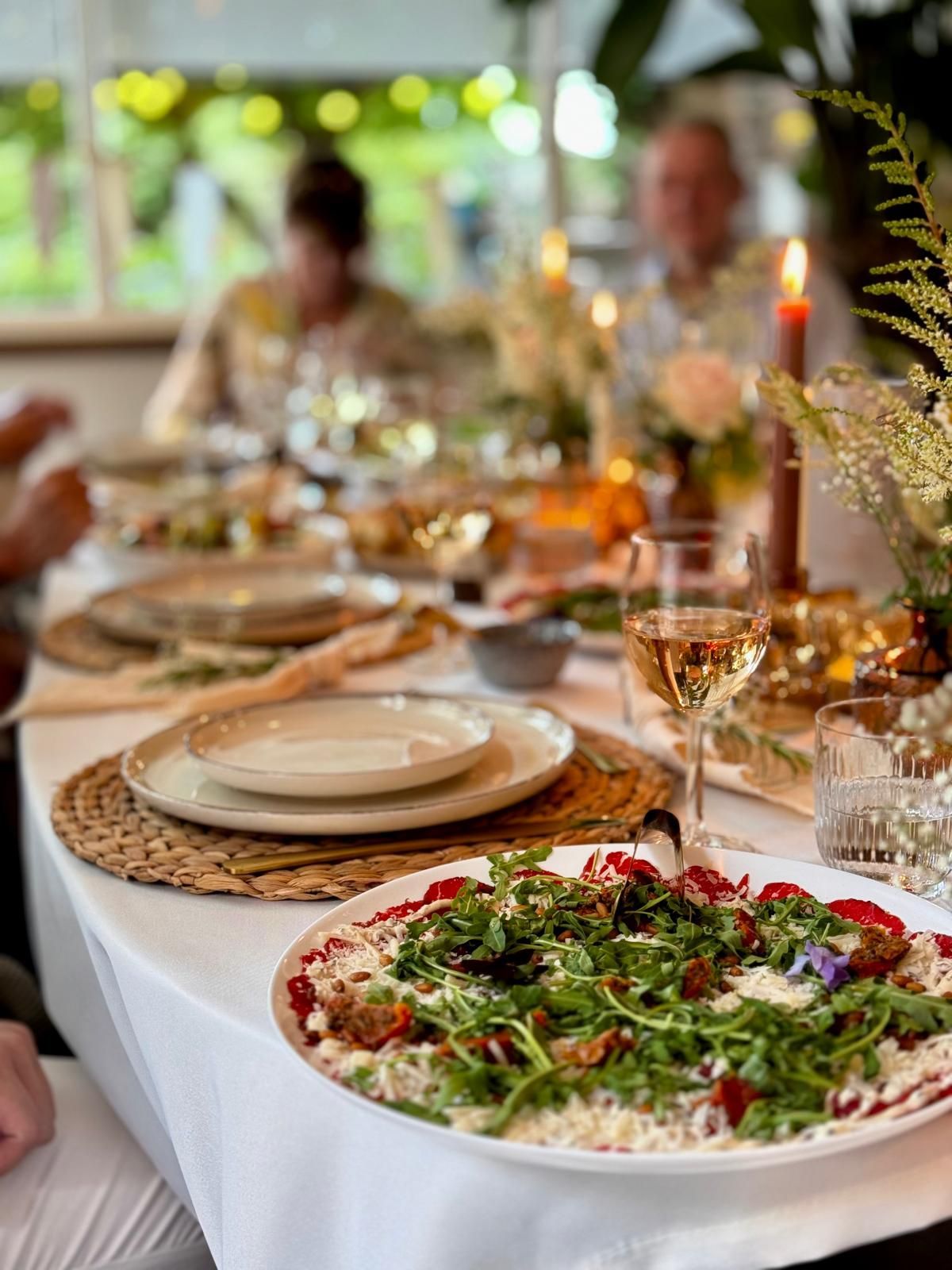 De tafel is gedekt voor een diner met borden, wijnglazen en een grote schaal salade met rucola en bessen.