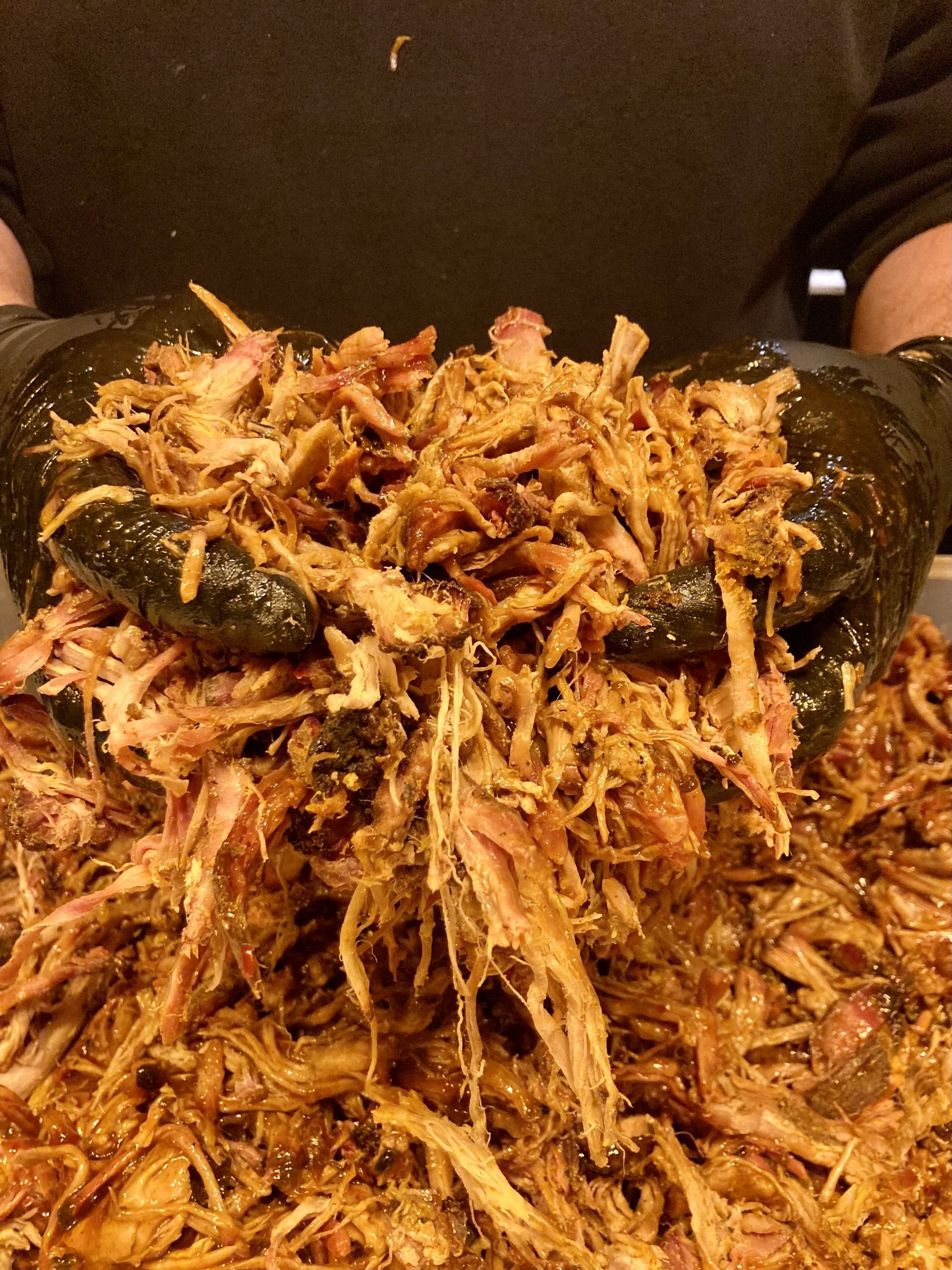 In zwarte handschoenen gehulde handen ligt een grote berg geraspte, met saus bedekte pulled pork.