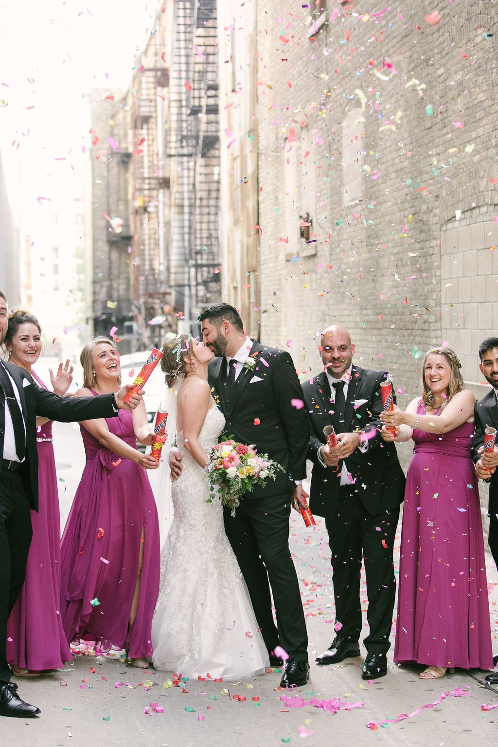 Wedding couple walking through confetti in a narrow stone alley, bridesmaids in purple dresses beside them