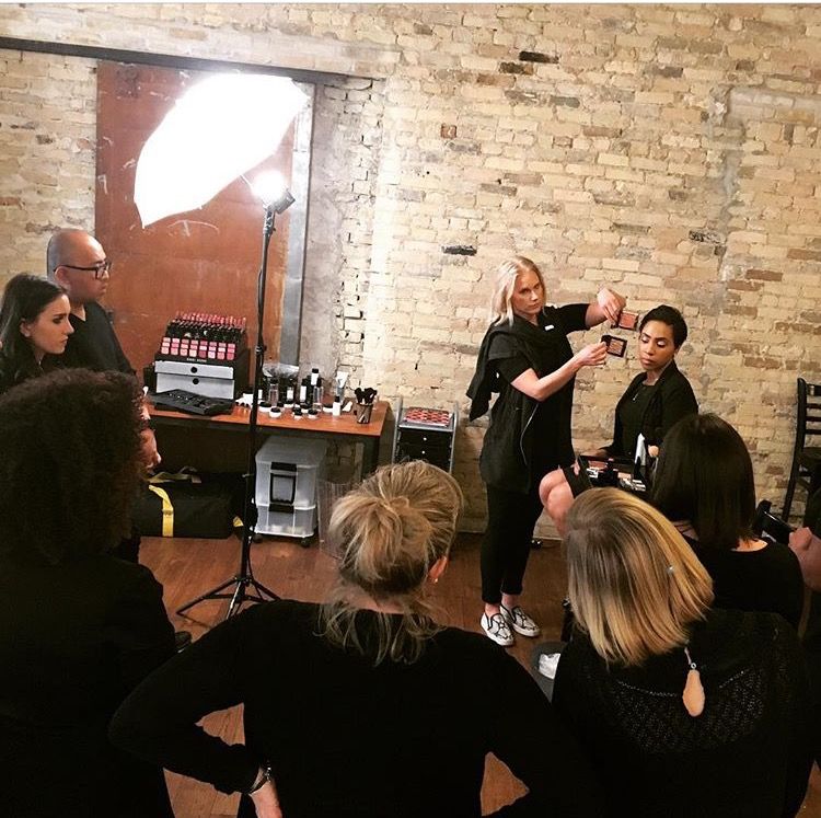Hair styling demonstration in a brick-walled room, with an audience watching a model and stylist.