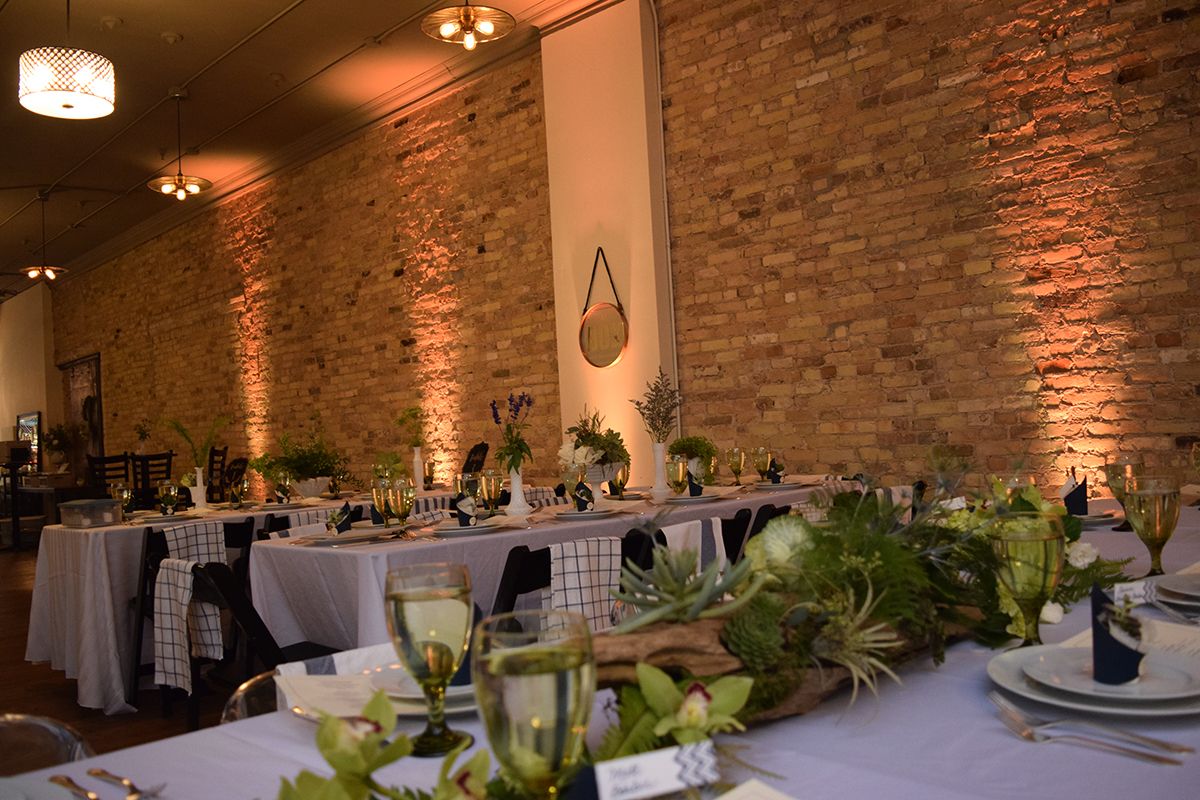 Elegant banquet hall with long white-draped tables, greenery centerpieces, and warm brick walls