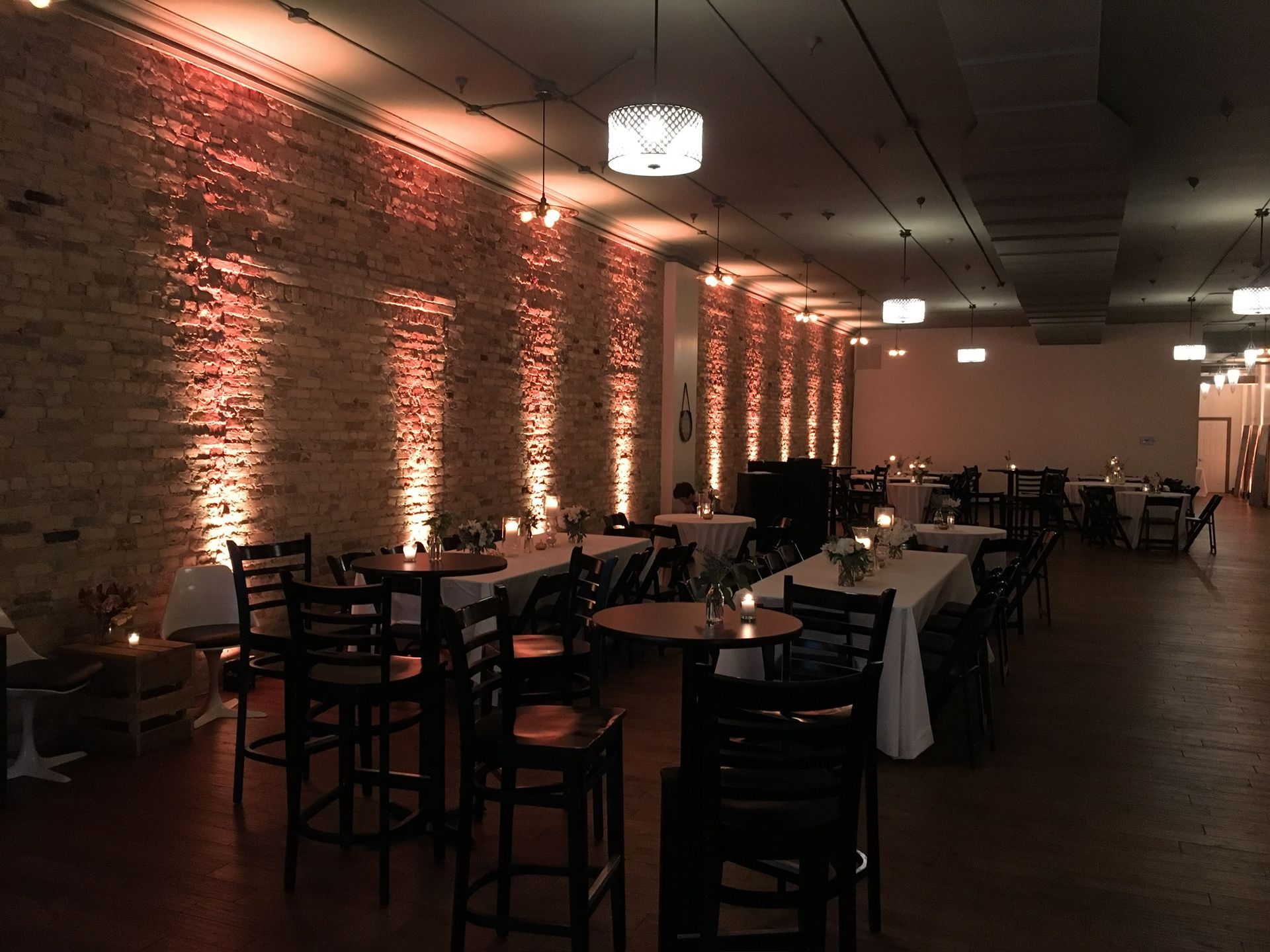 Dimly lit banquet hall with long tables, dark chairs, brick wall, and warm uplighting