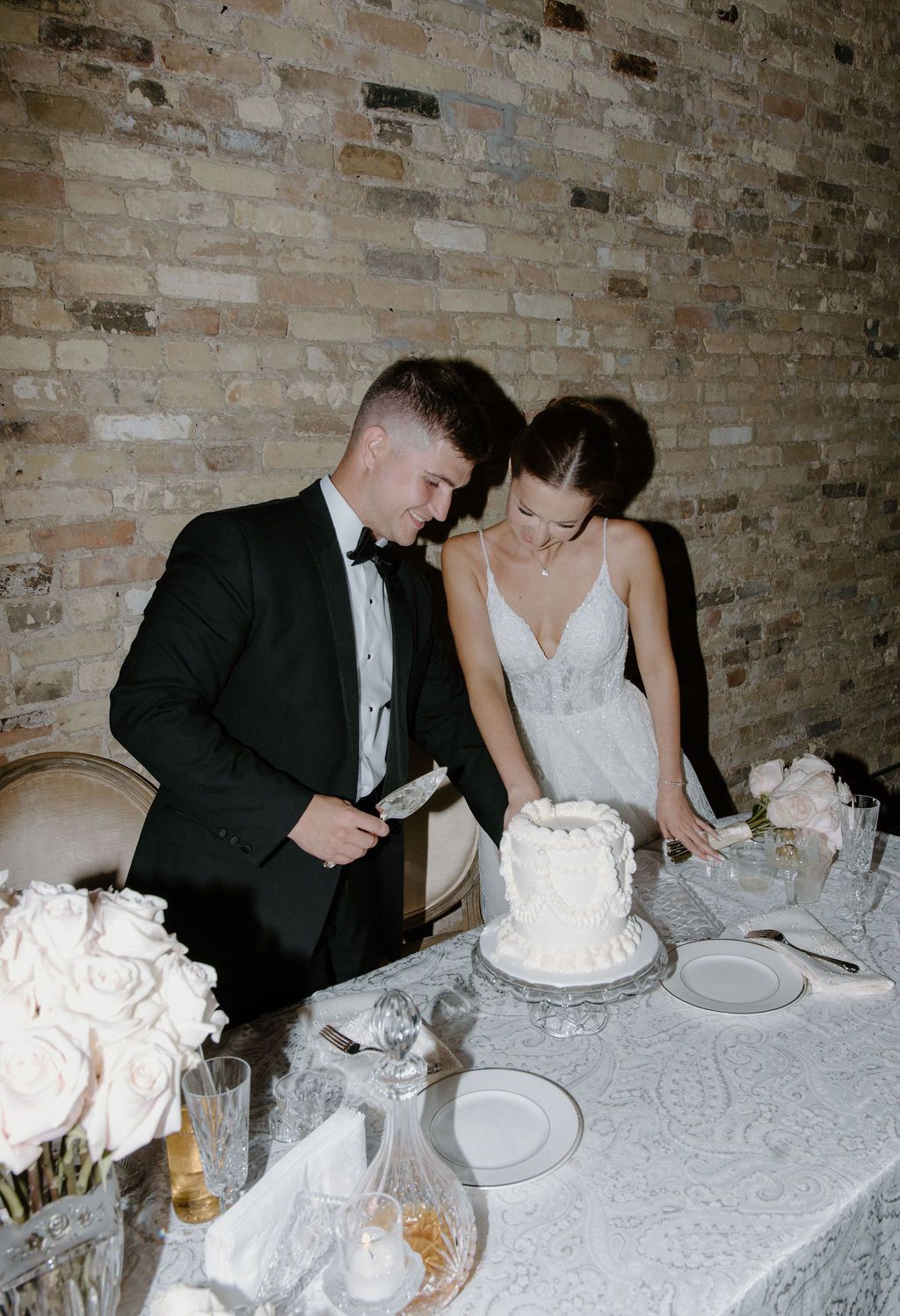 Wedding couple cutting a cake at a decorated reception table, with a brick wall backdrop