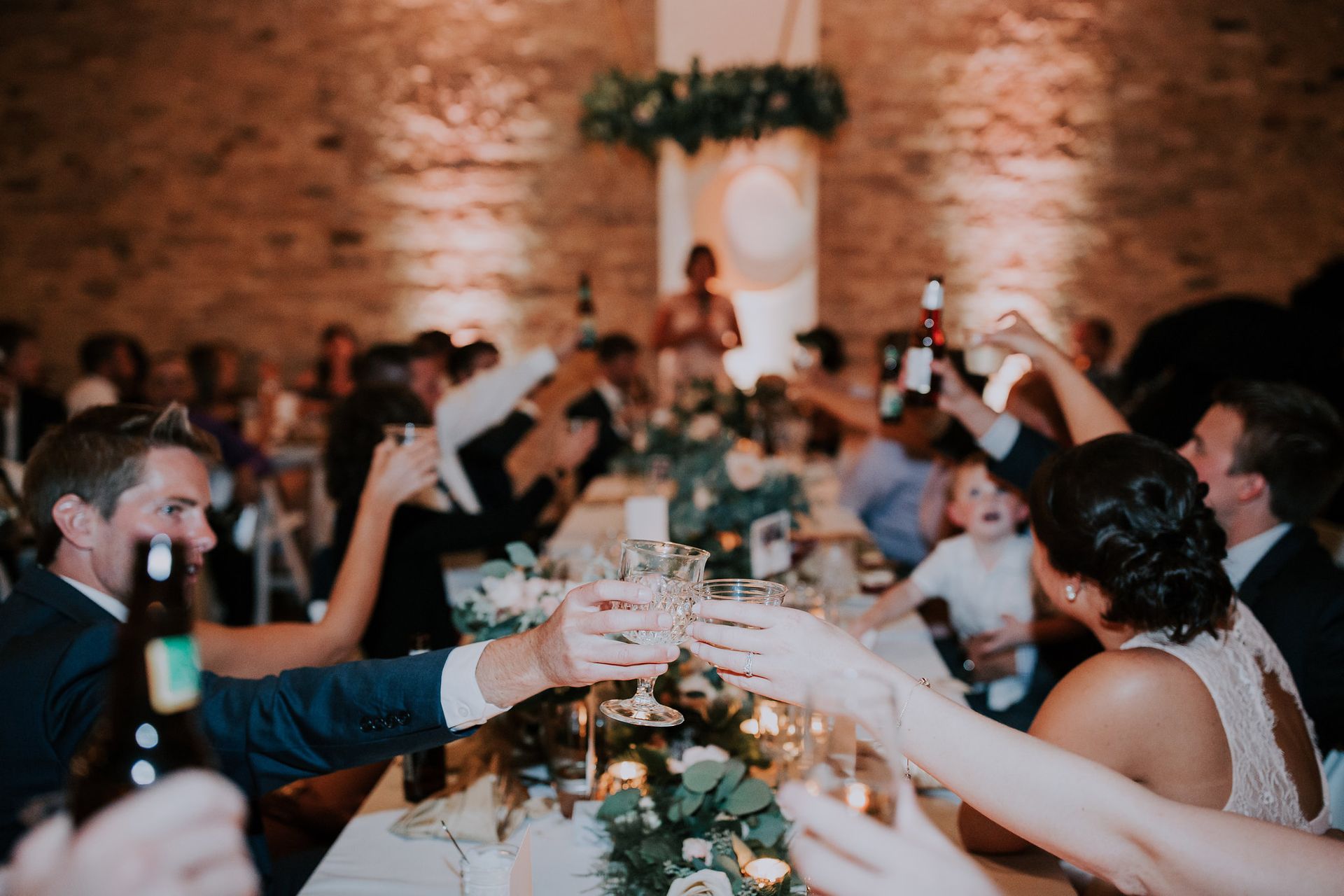 Guests toast at a long candlelit banquet table in a rustic brick-walled hall.