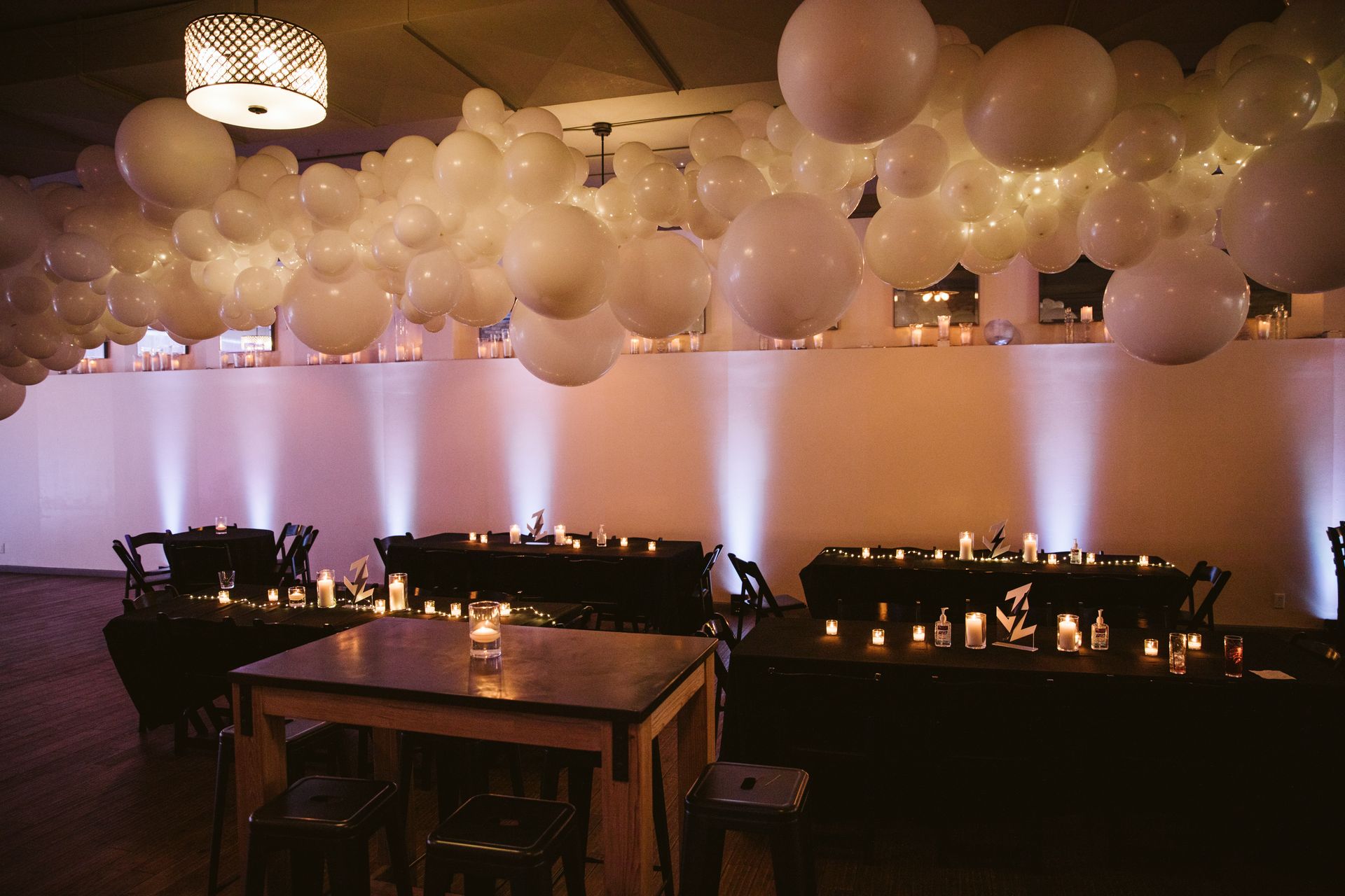 Dimly lit event space with white balloons overhead, candlelit tables, and a wooden table in the foreground