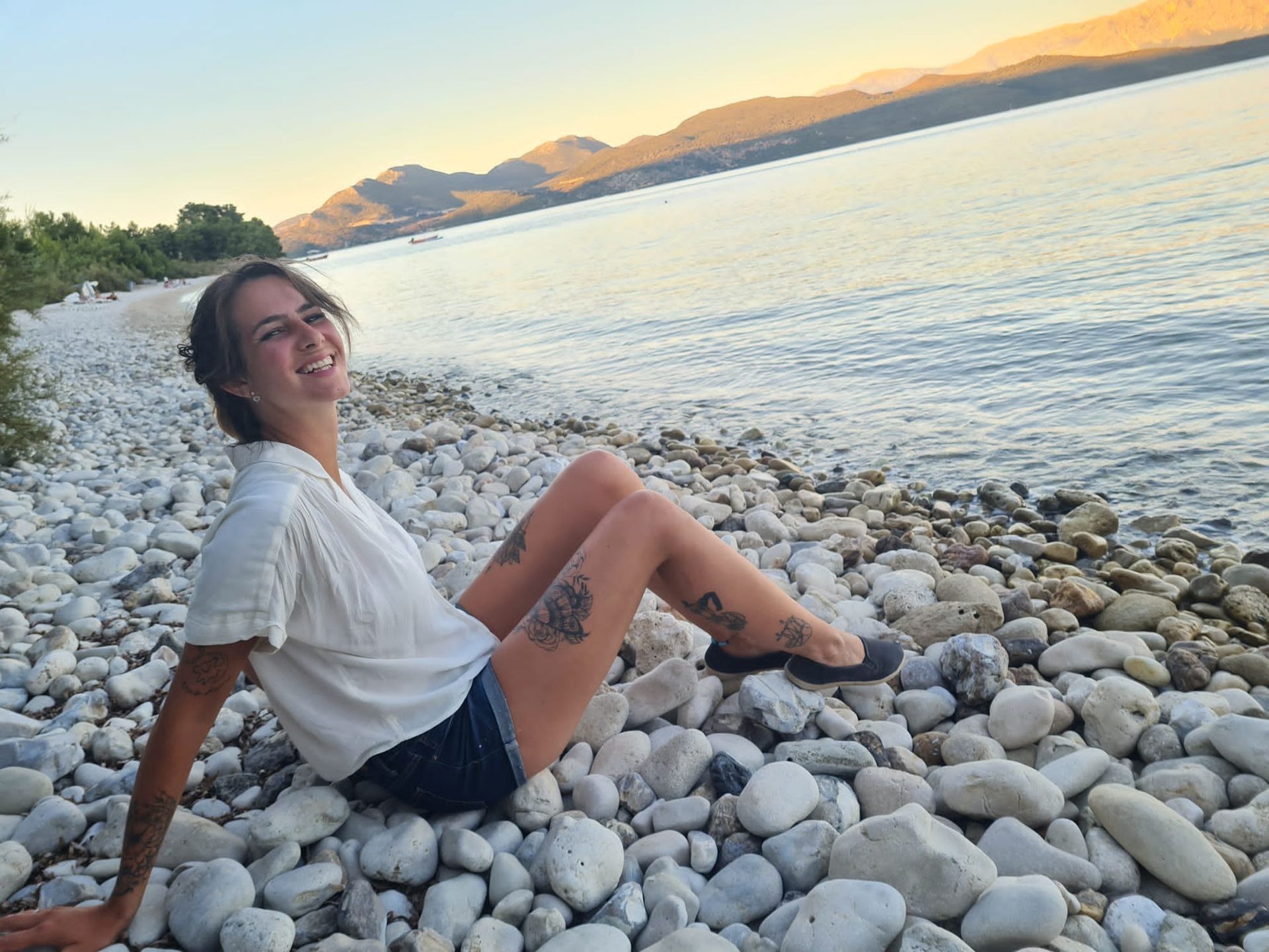 Claudia Bierbrauer Diaz sitzt lächeln auf einem Steinstrand am Meer, im Hintergrund sind Berge zu sehen.