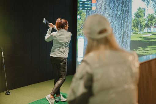 Woman swings a golf club in an indoor simulator, watched by another woman. Bright screen shows a golf course.