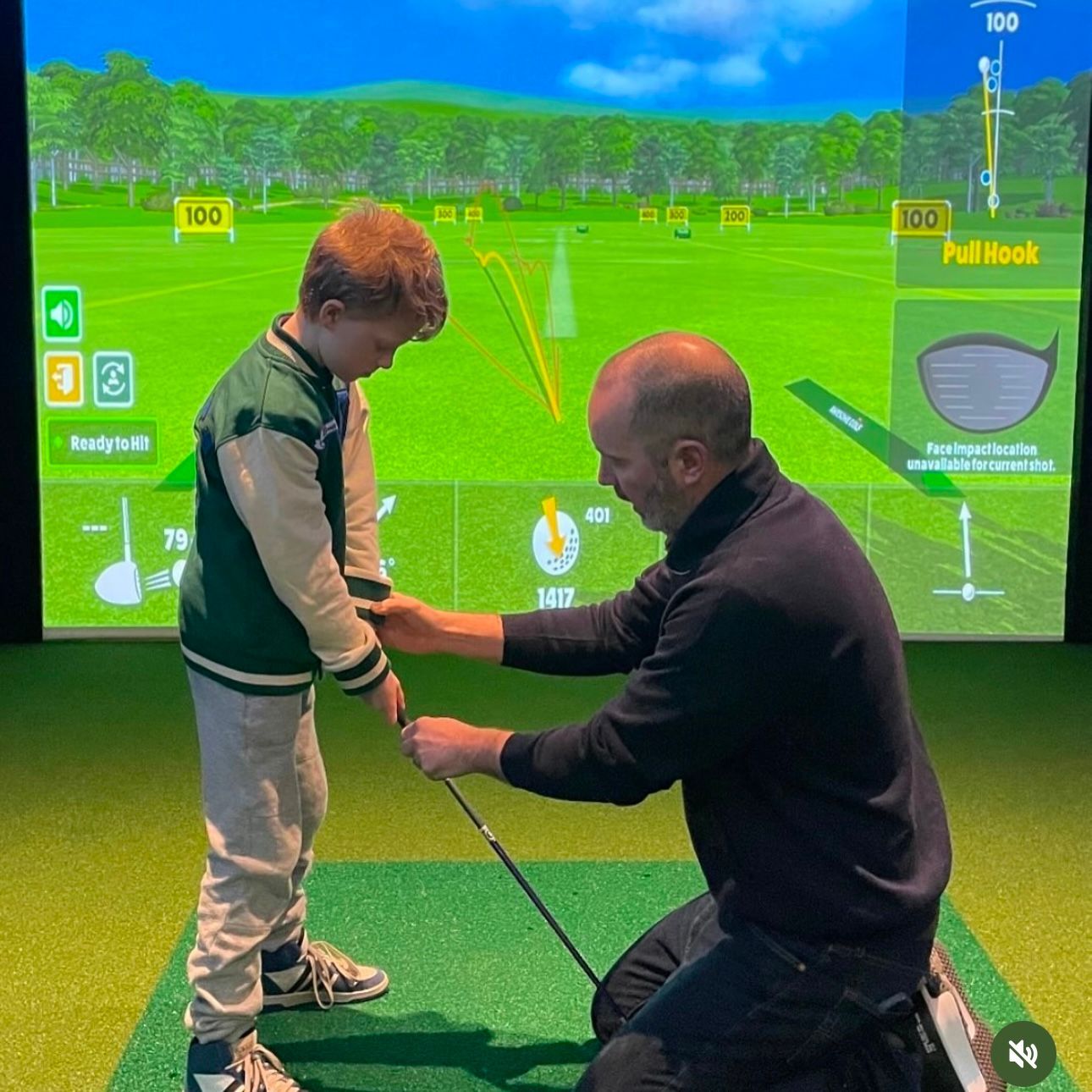 A coach helps a young boy with his golf swing at an indoor golf simulator. The boy is holding a club, and the screen shows a course.