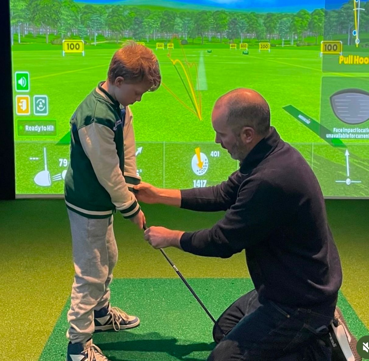 A coach helps a young boy with his golf swing at an indoor golf simulator. The boy is holding a club, and the screen shows a course.