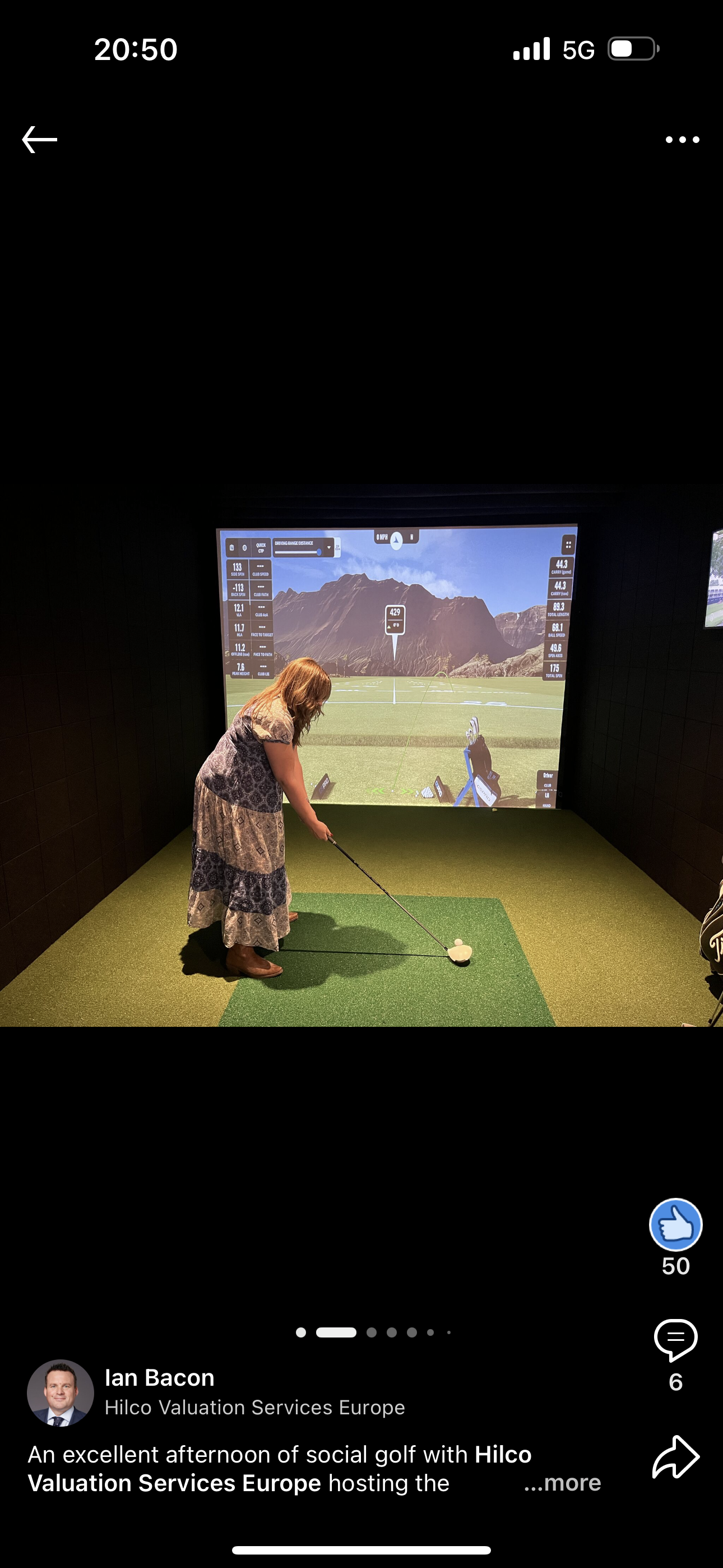 A woman in a patterned dress swings a golf club in an indoor golf simulator. A screen shows a golf course.