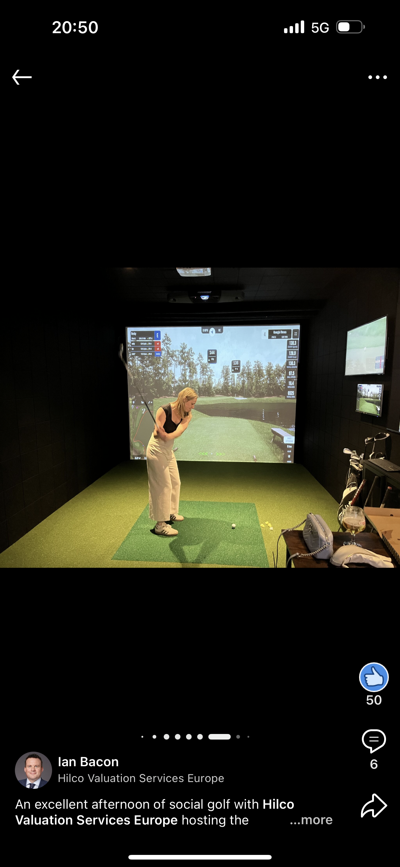 A person in a golf simulator wearing light-colored clothing prepares to swing a club. They are inside a dark room with a green hitting surface.