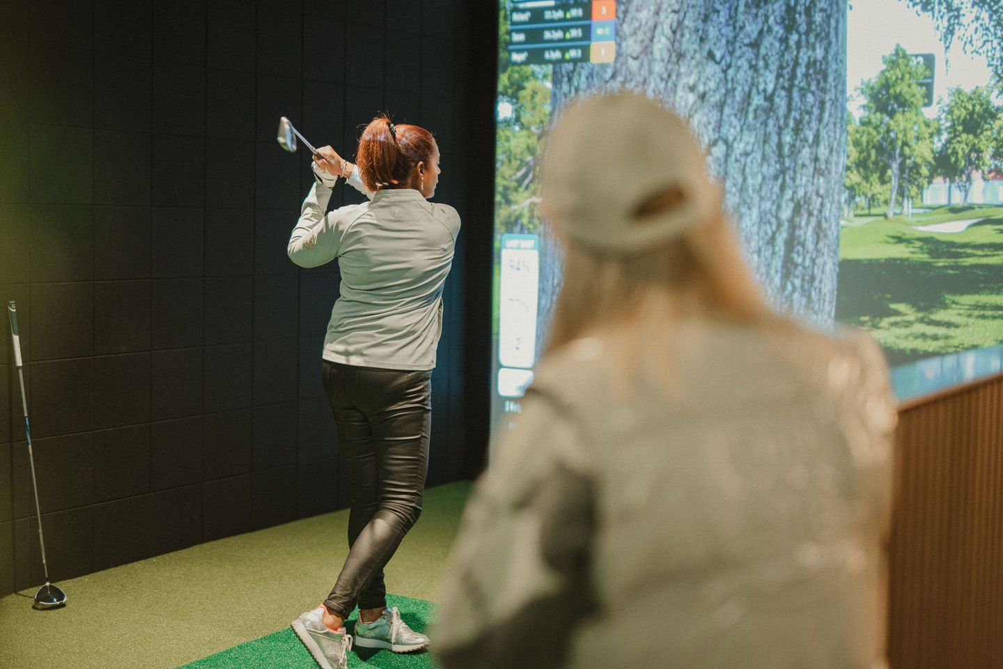 A woman in black pants and a light jacket swings a golf club indoors, viewed from behind. A second woman watches, wearing a cap.