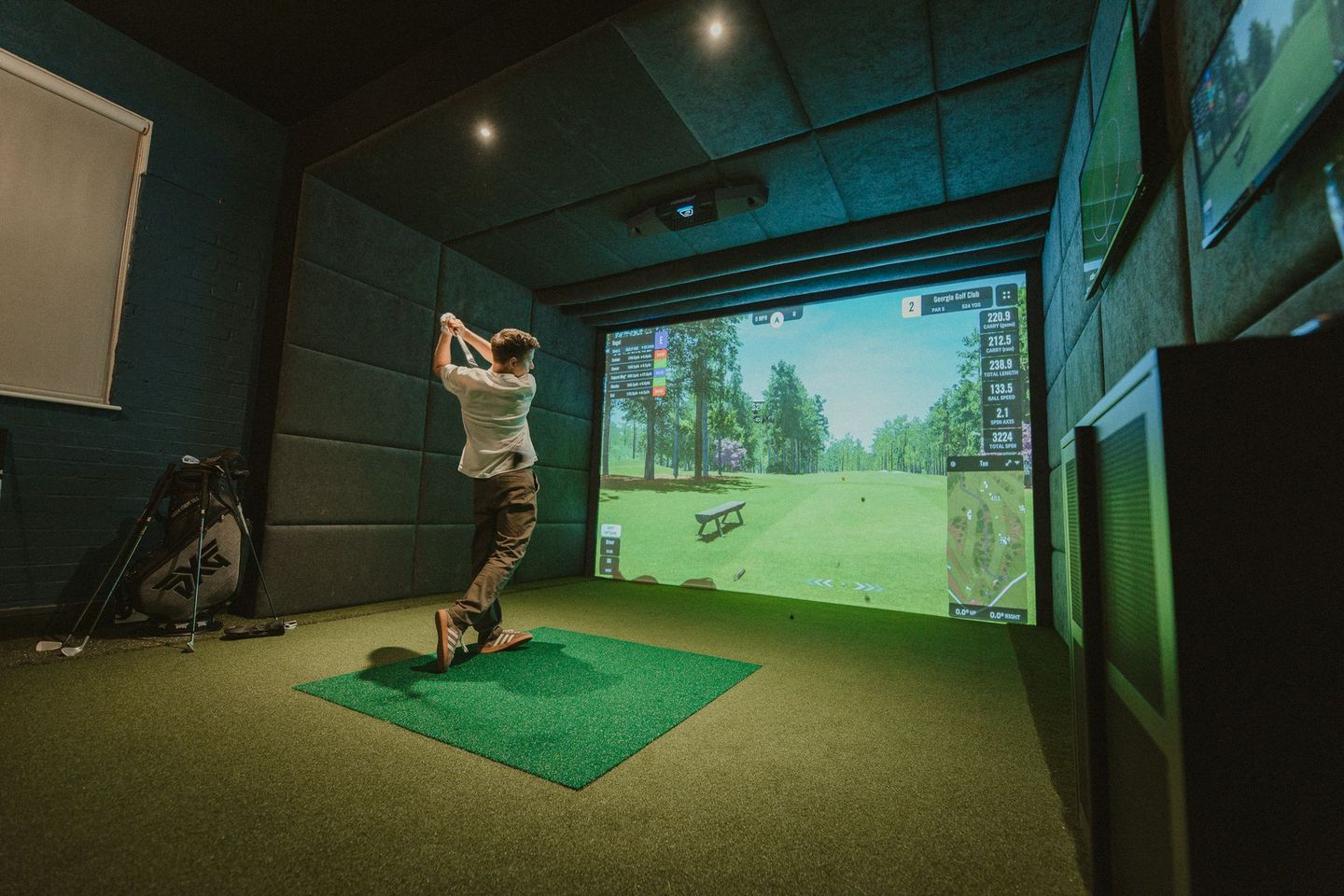 A person swings a golf club in a golf simulator. They are in a dark room with a green mat and a screen displaying a golf course.