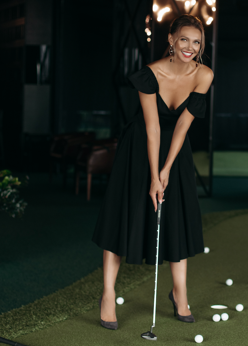 Woman in a black off-shoulder dress smiles while putting on a miniature golf course. Balls and clubs are scattered on the green turf.
