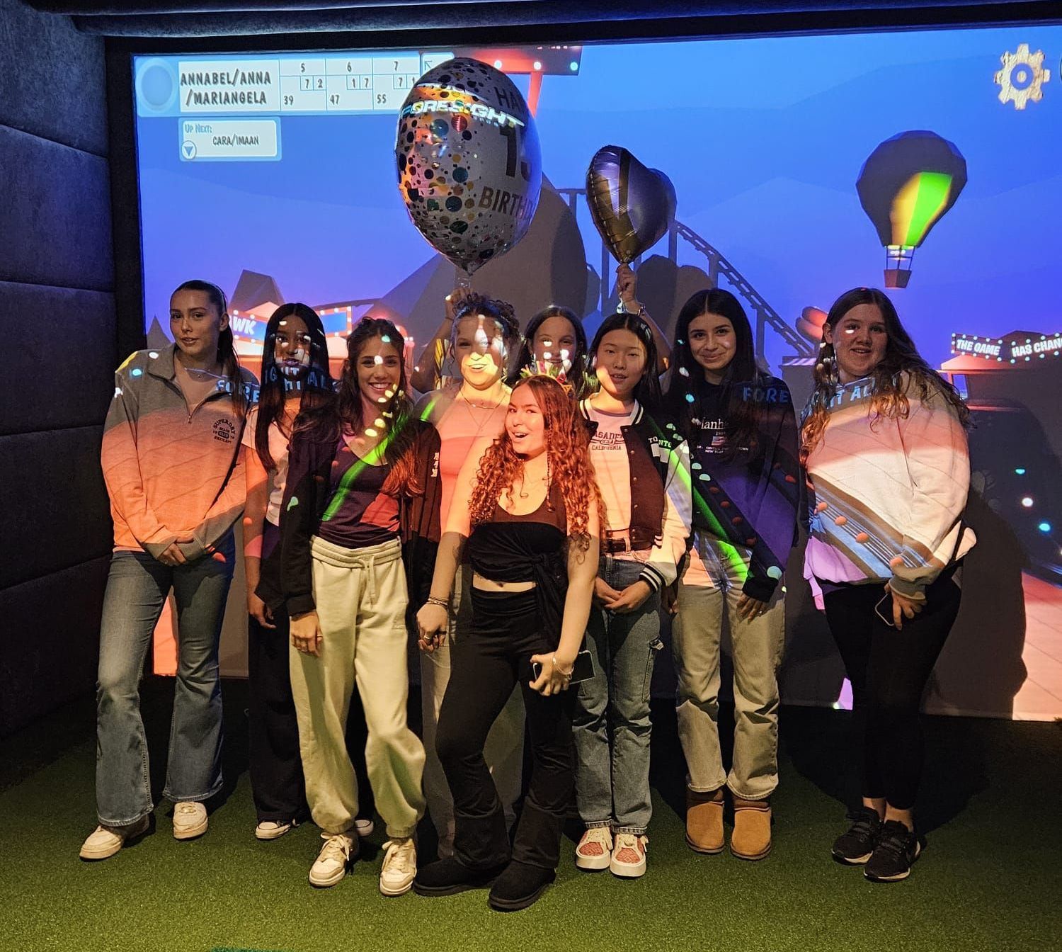 A group of young people poses in front of a virtual cityscape with balloons. The group smiles and has various outfits and face paint.