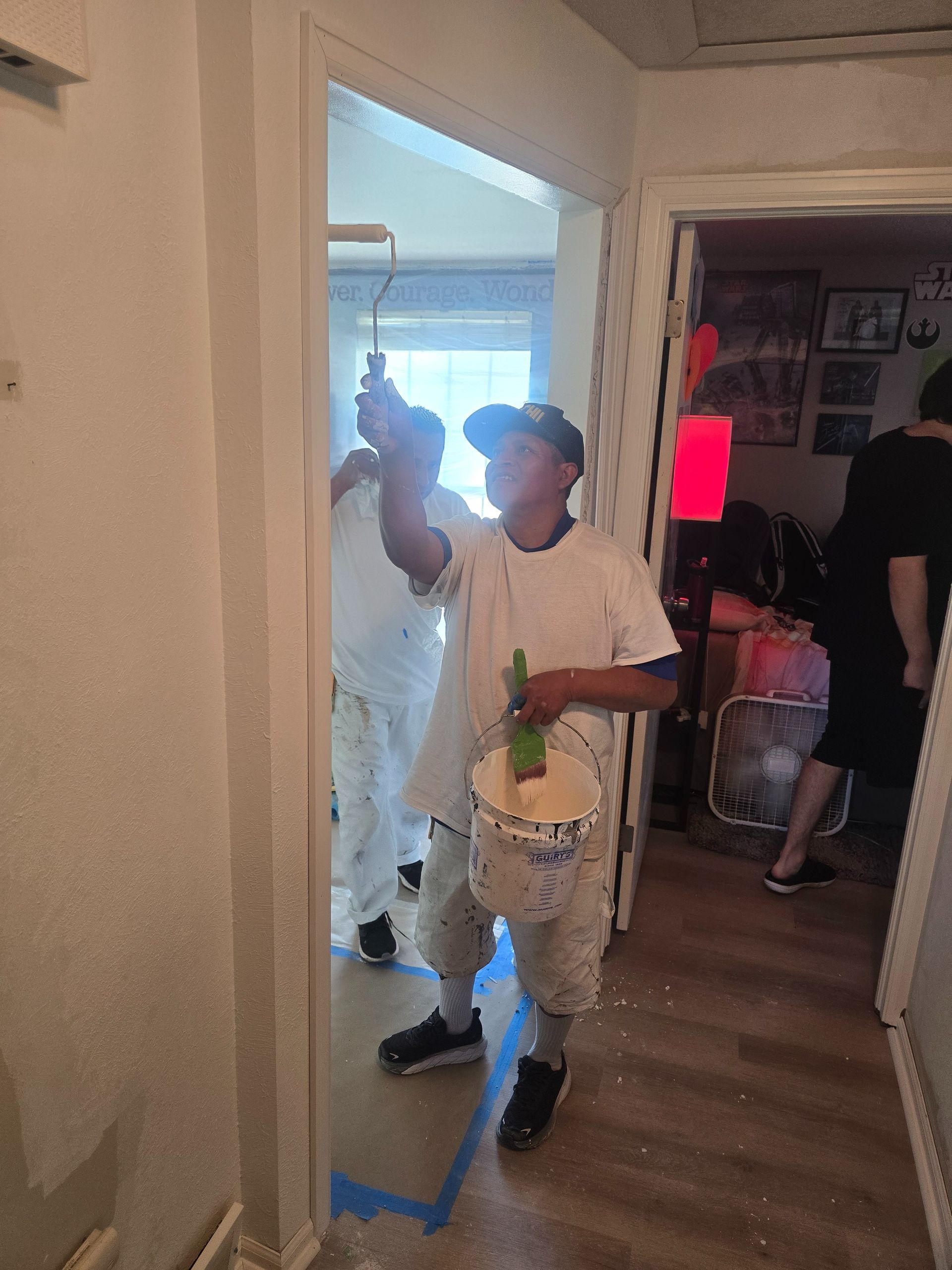 Two people painting a doorway. One rolls paint, the other holds a bucket. White paint, blue tape on floor.