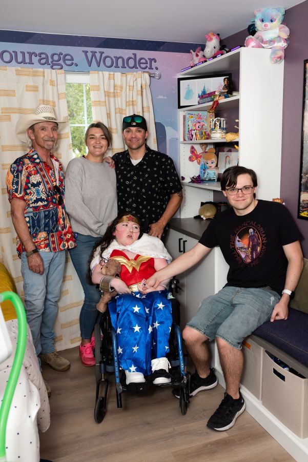 A group poses with a child in a Wonder Woman costume, inside a colorful bedroom.
