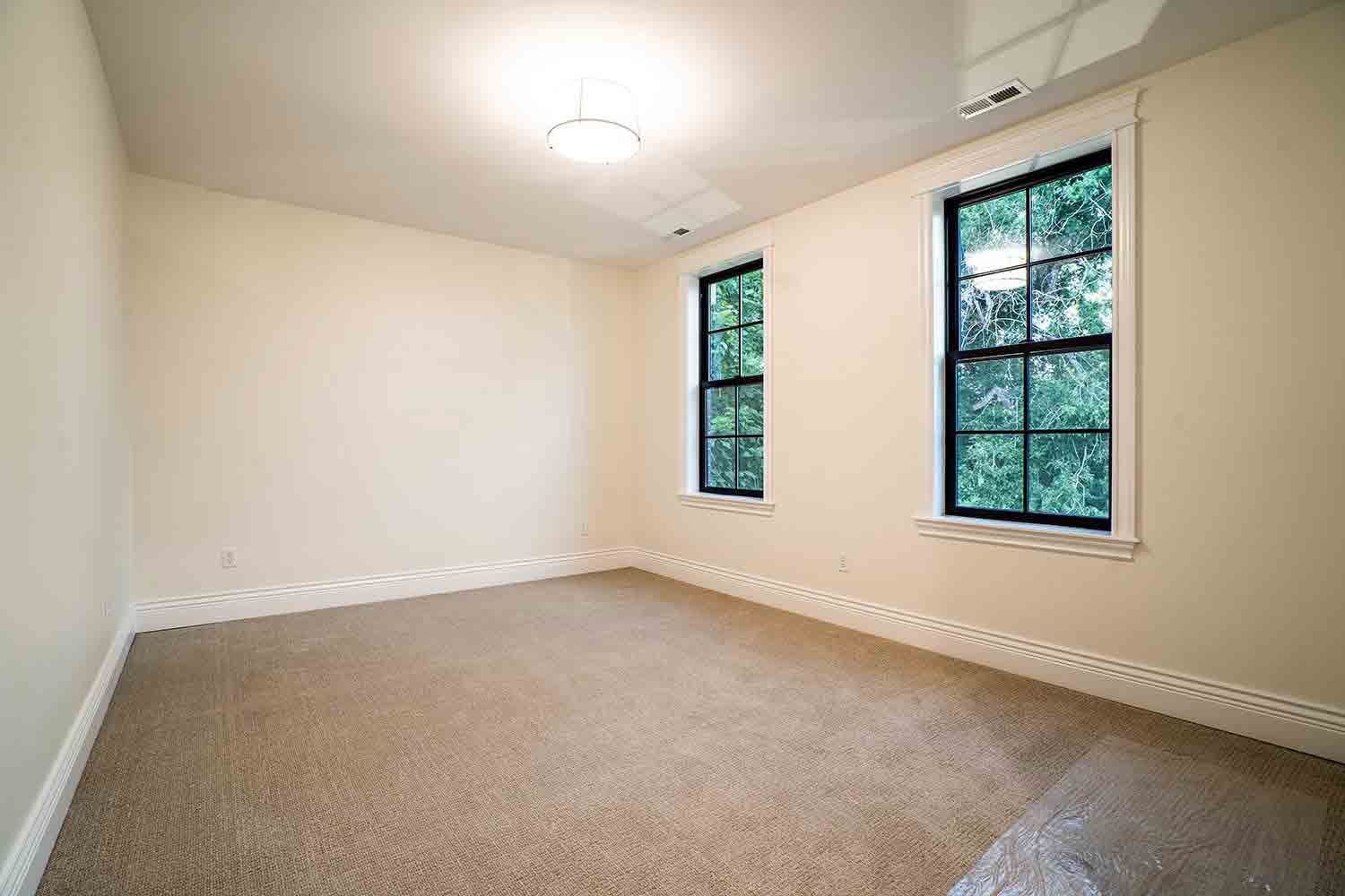 An empty room with beige walls, tan carpeting, white trim, and two windows looking out onto green trees.
