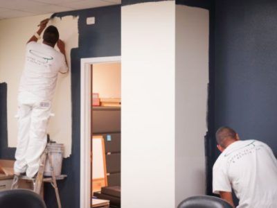 A man on a ladder is painting a wall while another man looks on