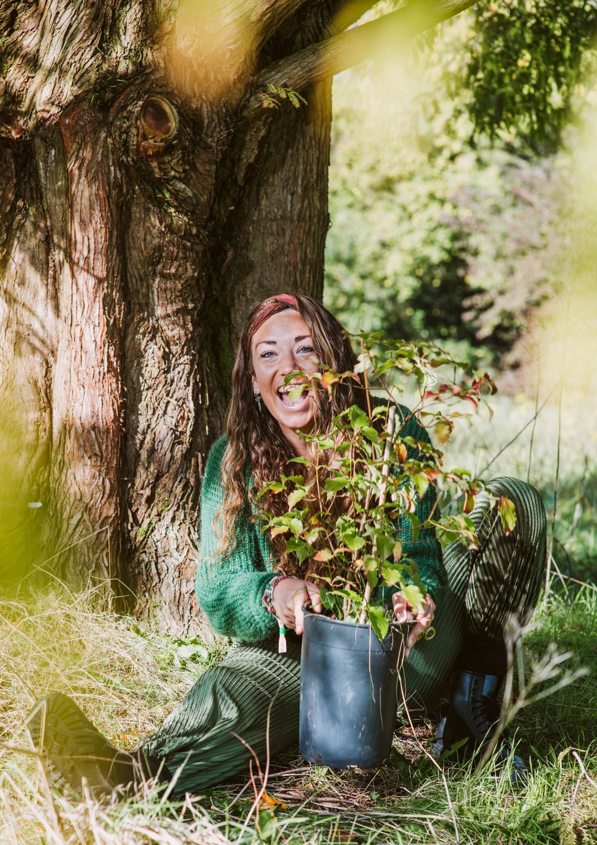 Een vrouw met een bruine hoed en een roestbruine jas lacht buiten, met lang golvend haar en groen op de achtergrond.