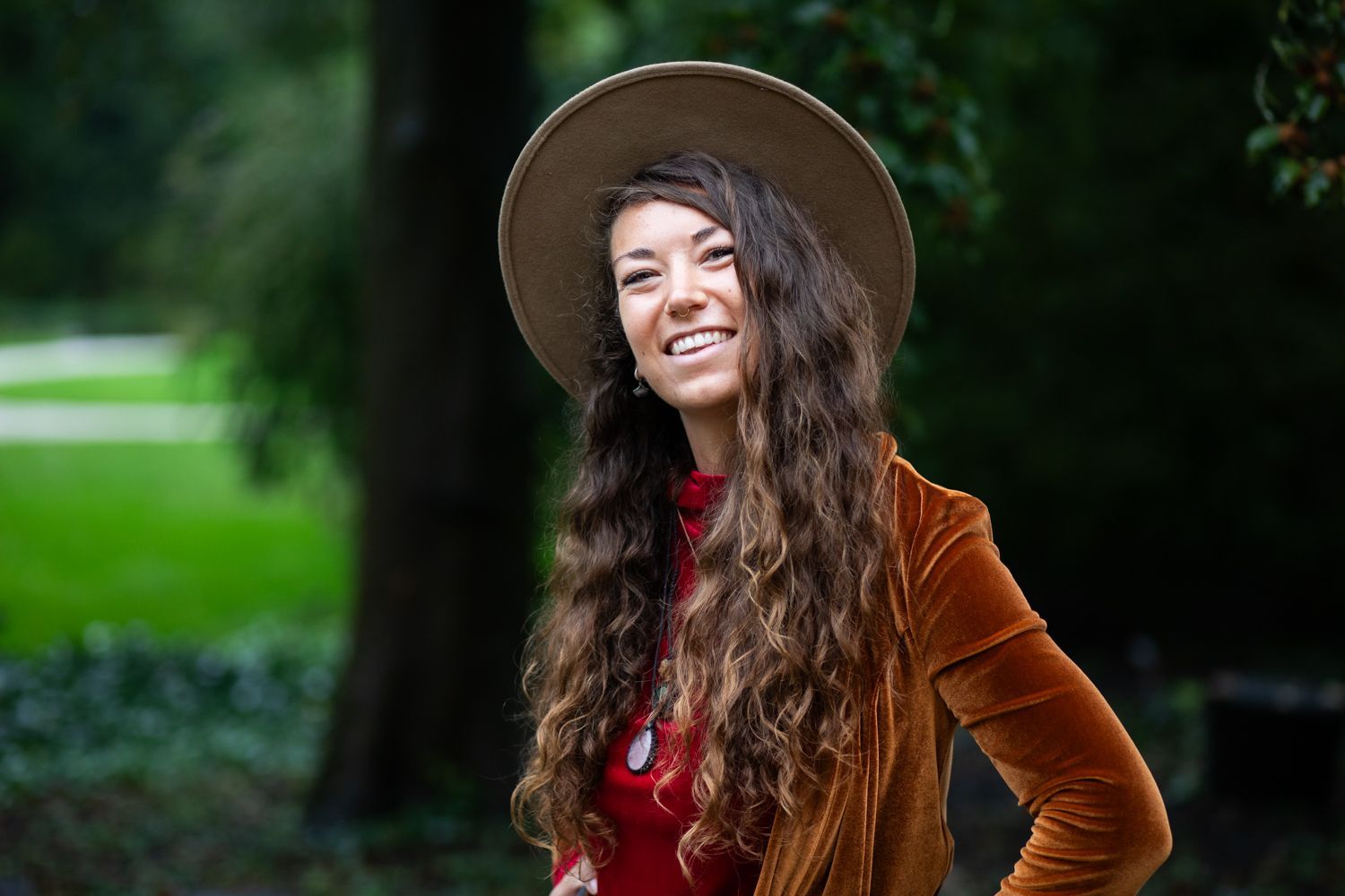 Een vrouw met hoed en oranje jas lacht buiten, met lang golvend haar en een groene achtergrond.