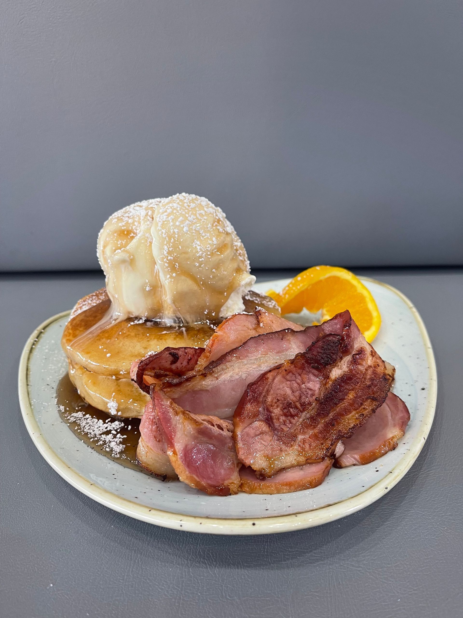 Pancakes with a scoop of ice cream, bacon, and an orange slice on a plate, sprinkled with powdered sugar.