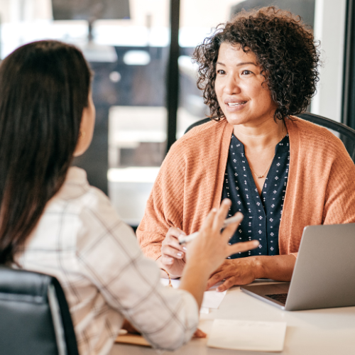 Two women are sitting at a table with a laptop and talking to each other.