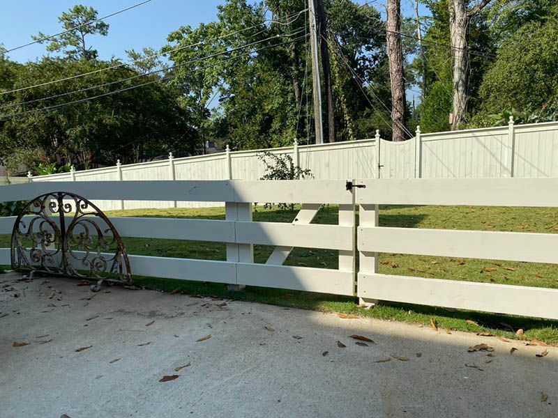 A white fence with a gate and a decorative iron piece. The fence separates a lawn from the background trees.