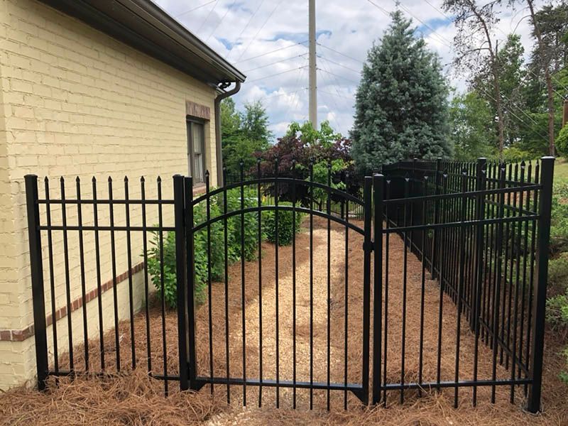 Black metal fence with arched gate, bordering a pathway lined with bushes, next to a beige brick building.
