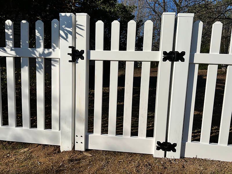 White picket fence with a matching gate, secured by black hinges and a latch, set on brown ground.