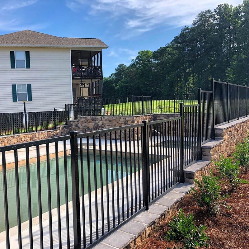 Black metal fence surrounds a swimming pool and stone retaining wall next to a two-story white house with a screened porch; trees in the background.