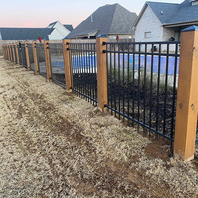 A black metal fence with wooden posts surrounds a pool area in a residential yard. Brown grass and houses are in the background.