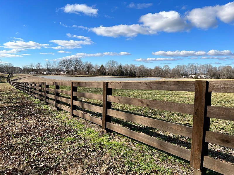 Wooden fence bordering a field, with a pond and trees in the background under a blue sky with fluffy clouds.
