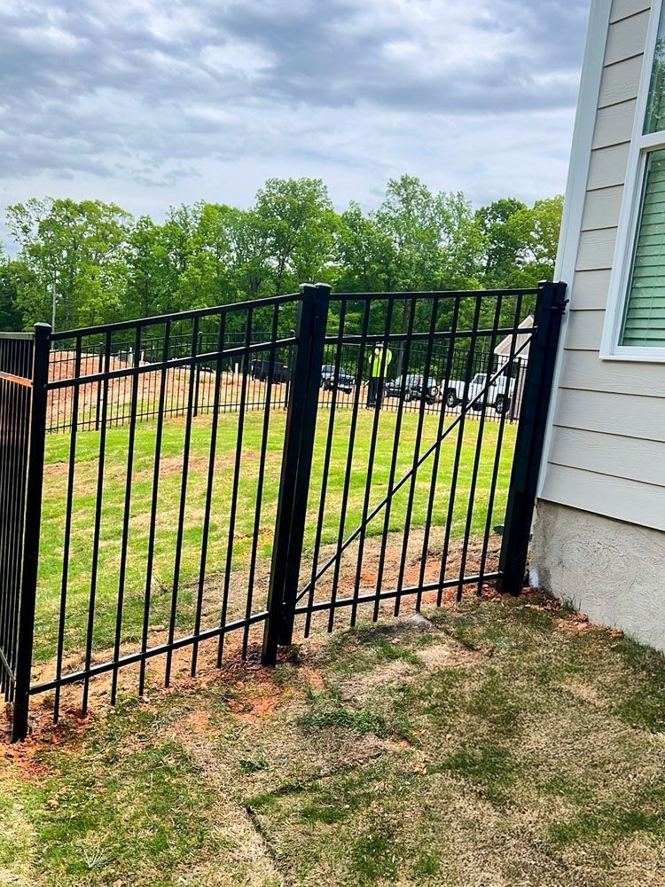 Black metal fence angled in a yard, attached to a building's beige siding. Green grass and trees in the background.
