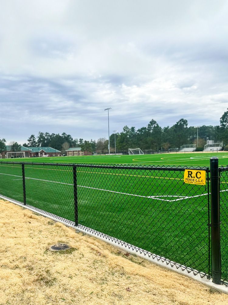 Black chain-link fence surrounding a green sports field under a cloudy sky. A yellow sign reads 