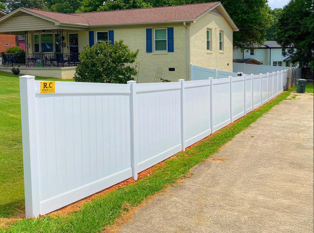 White vinyl fence along a paved path, beside a grassy lawn and two-story beige house with blue shutters.