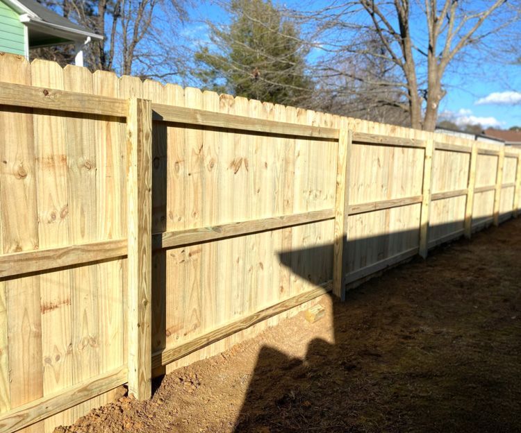 Wooden privacy fence in a backyard, constructed with vertical boards and framed posts. The fence is new, and unpainted, on a sunny day.