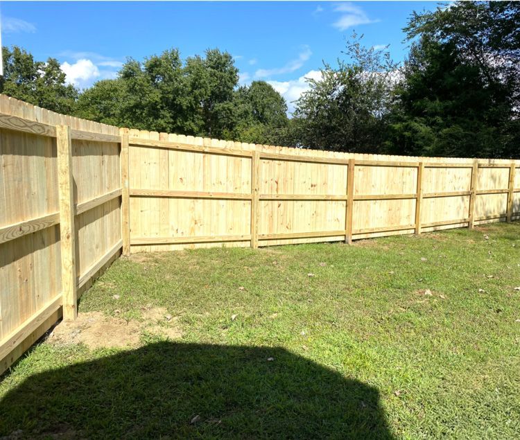 A wooden fence in a grassy backyard, under a blue sky with trees in the background.