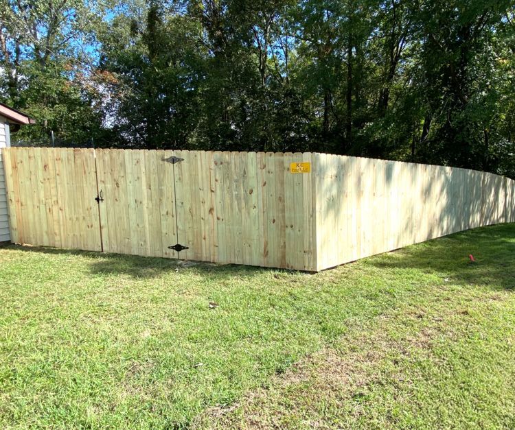 A wooden fence with a gate in a grassy yard, trees in the background, under a blue sky.