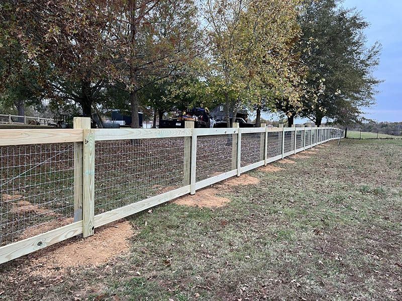 Wooden fence with wire mesh panels, in a grassy field, with trees and a vehicle in the background.