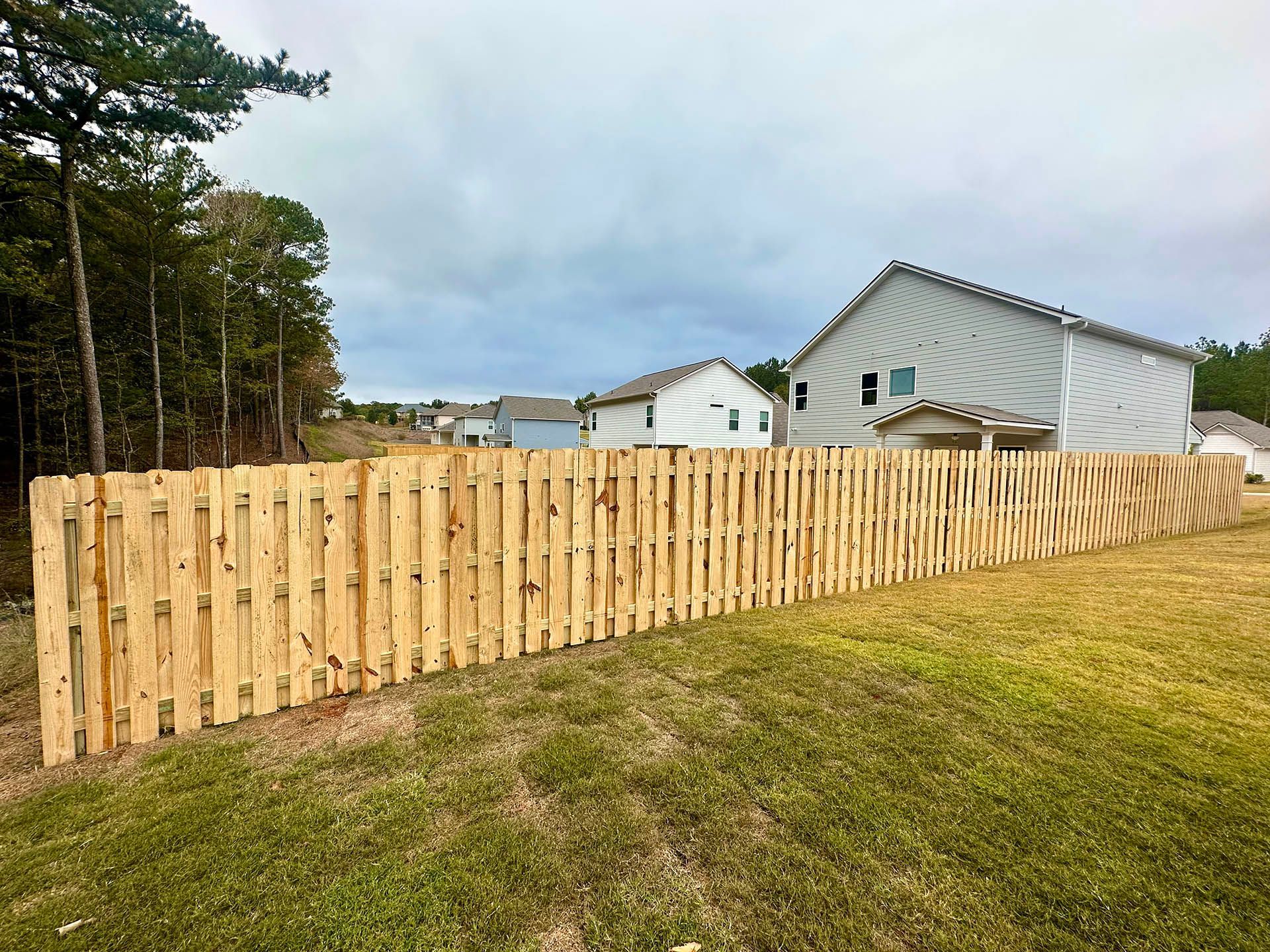 Wooden fence in a grassy yard, with houses and trees in the background under a cloudy sky.