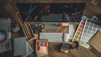 Overhead view of a cluttered desk with keyboard, mouse, coffee mug, notebooks, and color swatches on wood