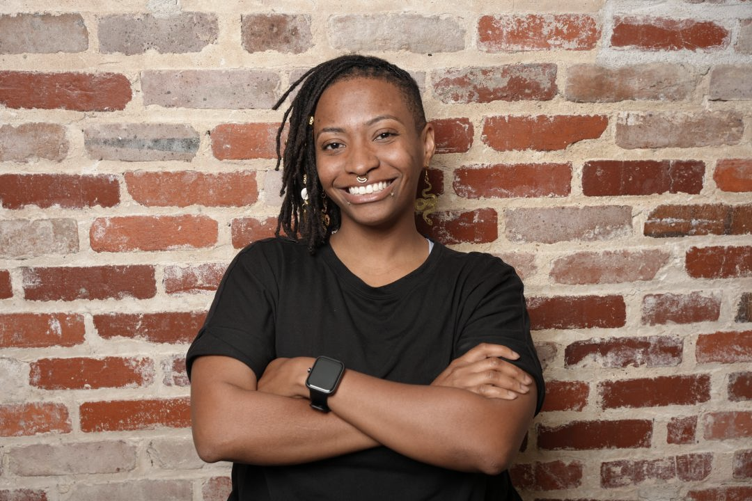 Smiling person with crossed arms in a black shirt against a brick wall