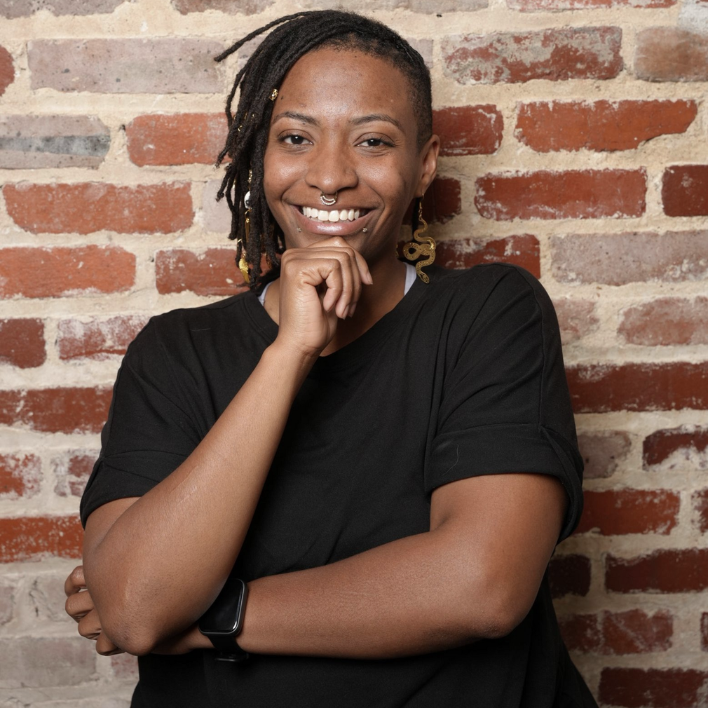 Smiling person with crossed arms in a black shirt against a brick wall