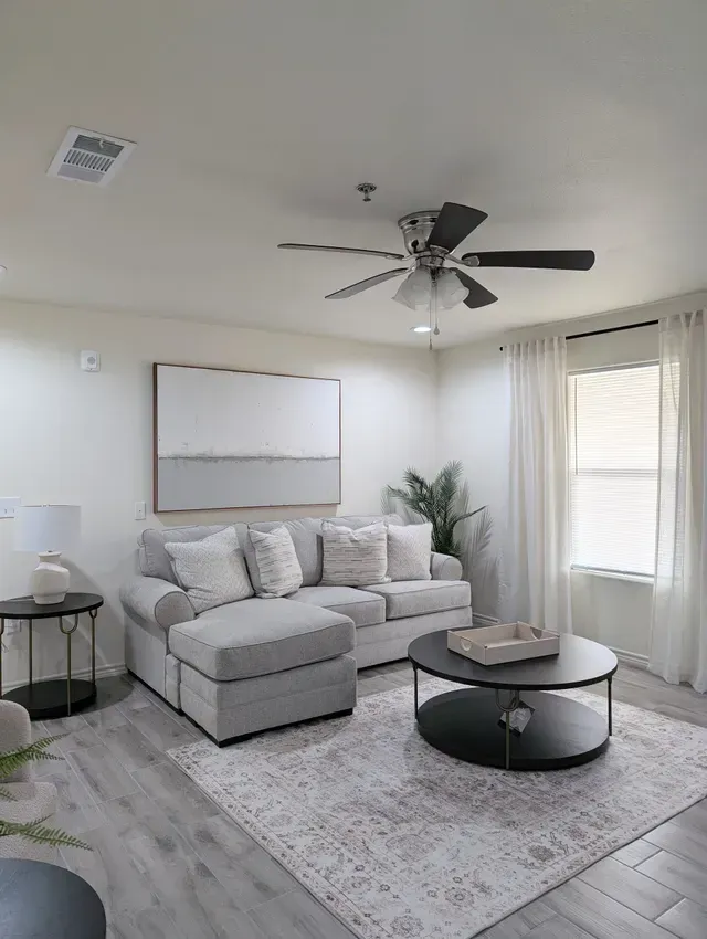 Bright living room with gray sectional, round coffee table, wall art, fan, and window with curtains